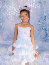 A young girl wearing a light blue and pink Barbie x Tutu du Monde tutu dress with a matching headband and tiara, standing in front of a snowy, icy backdrop with lights.