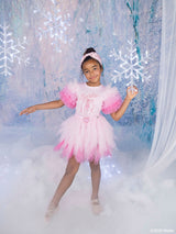 A young girl posing in a pink Barbie tutu skirt & top with fluffy pink sleeves, set against a magical winter-themed backdrop with glowing snowflakes and a snowy floor.