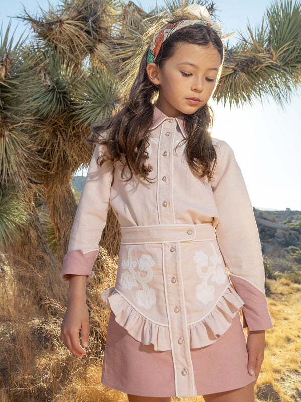 Young girl in a pink outfit standing in a natural setting with plants and sky.